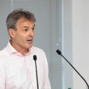 Man in salmon pink shirt speaking at podium.