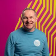 Man in blue jumper in front of colourful pink and yellow wall.