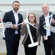 Two men and a woman holding models of wind turbines on a beach.
