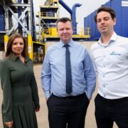 Woman in green dress beside two men in business attire in front of recycling machines.