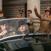 Man and woman in vintage sports car with people in background wearing long coats and hats.