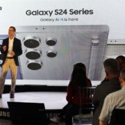 Man speaking on stage with image of a Samsung Galaxy flip phone in background.