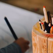 Child drawing beside a container full of coloured pencils.