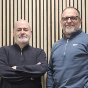 Two men in front of a wooden wall.