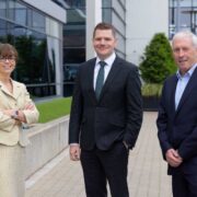 Woman in cream suit beside 2 men in dark suits.