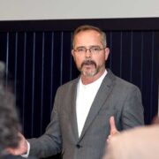 Man in grey jacket speaking at an event.