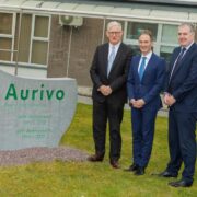 Three people in business attire stand beside a large engraved stone sign on a grassy area outside a building. The sign displays the Aurivo logo and text marking the 20th and 50th anniversaries of the company&rsquo;s dairy ingredients operations.