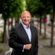 A person wearing a dark suit jacket and light shirt stands outdoors on a tree‑lined pathway with evenly spaced trees and a paved walkway in the background.