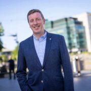 A man wearing a blue blazer and light blue shirt standing outdoors in an urban setting with modern glass buildings, trees, and a pedestrian walkway in the background.