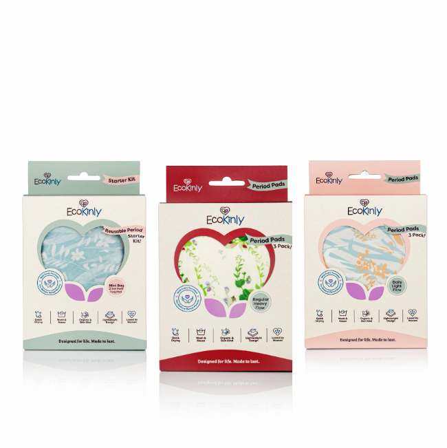 Three EcoKinly product boxes displayed side by side, each with a heart‑shaped window showing reusable period pads in different patterns. The left box features a blue starter kit, the centre box shows a regular pack with a green botanical pattern, and the right box displays a pink‑themed pack with a blue pattern. The packaging highlights features such as absorbency, comfort, and reusable design.