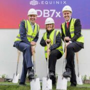 Three individuals wearing safety helmets and high‑visibility vests pose outdoors with ceremonial shovels during a groundbreaking event in front of a large Equinix DB7x project sign. They are standing on grass with small white planters arranged behind them and additional shovels placed upright along the wall.