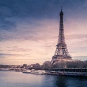 A wide view of the Eiffel Tower beside the River Seine during sunset, with boats moored along the riverbank and soft pink and blue clouds in the sky.