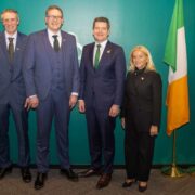 Four men and a woman in New York standing before a green wall alongside flags of Ireland, EU and USA.
