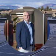 Solar panels installed on a rooftop with a circular inset showing a person in business attire standing in a modern office hallway, with a town and mountains visible in the background.