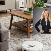 A modern living room with a patterned rug, grey sofas and a wooden coffee table with books and a plant, with a smart robot vacuum on the floor and an overlaid portrait of a person standing with hands on hips wearing a dark blazer.