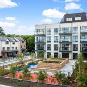 Modern residential development featuring a multi‑storey apartment building and adjoining townhouses, overlooking landscaped communal gardens with trees, pathways, and raised planting beds under a bright blue sky.