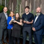 Group of five adults standing indoors at an AxisBIC Venture Academy event, with two people holding glass awards, photographed against a leafy mural backdrop at the River Lee Hotel in Cork.