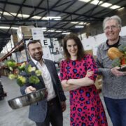 Three people stand inside a food redistribution warehouse, with pallet racking visible in the background. One holds a metal bowl of fresh herbs, another stands with arms folded, and a third holds a crate containing bread and vegetables, illustrating surplus food redistribution.
