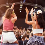 Two people seated on others&rsquo; shoulders clap above their heads in front of a large outdoor festival stage, with a dense crowd below and stage lighting and rigging visible in the background.