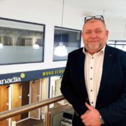 Person wearing a dark blazer and light shirt stands by a glass railing inside a wood flooring showroom, with hanging lights and &ldquo;Canadia Wood Flooring&rdquo; display panels visible below.