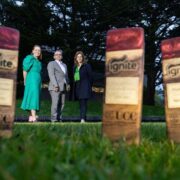 Ignite awards wooden trophies in the foreground with three participants standing on a grassy lawn on the UCC campus during the IGNITE Spring Awards event.