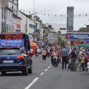 Crowds gather on a main street in Longford town near the finish line of the Longford Marathon, with timing clocks, race vehicles, banners, and bunting visible among shops and buildings.