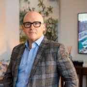 Portrait of Dominick Miciotta indoors, wearing glasses, a check blazer and blue shirt, standing in a modern living space with artwork and furnishings in the background.