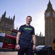 Man wearing a black TRYKA T‑shirt stands in the foreground on a London street, with the Elizabeth Tower (Big Ben), the Houses of Parliament, traffic, and a red double‑decker bus visible in the background under a clear sky.