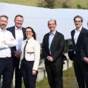 Six people standing outdoors beside rows of ground‑mounted solar panels at an industrial site, with metal panel structures in the background and greenery beyond, photographed during daylight.