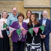 Eight adults stand outdoors in front of a modern brick building, with two people at the centre holding a knitted bunting that spells &ldquo;AGEWELL&rdquo; in colourful letters. The group is arranged in a straight line, dressed in smart-casual business attire, with landscaped greenery visible in the foreground and windows behind them.