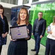 Four people standing in a modern office corridor with glass meeting rooms. One person in the foreground holds a tablet displaying a &ldquo;Benefits Finder&rdquo; webpage, while another to the right holds a smartphone showing the same service. The remaining two people stand slightly behind, facing the camera, with green and blue glass walls visible in the background.