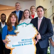 A group of eight people stand together indoors holding a large cut‑out sign shaped like the island of Ireland with the text &ldquo;All Island Bioeconomy Summit & Awards.&rdquo; Minister Martin Heydon is at the front right in a suit and pink tie. The group includes men and women in professional attire, smiling towards the camera. The background shows a staircase and modern interior setting.