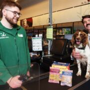 A Maxi Zoo store checkout counter displaying Butternut Box fresh dog food packs, with a store staff member, a shopper holding a dog on the counter, and retail shelves visible in the background.