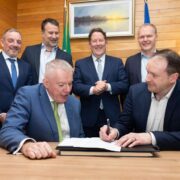 Group of business and government representatives seated and standing around a table as a contract is signed, marking an agreement for a solar carport project at Cork Airport, with Irish flags visible in the background.