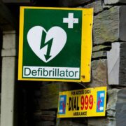 Green and yellow defibrillator sign with a white heart and lightning bolt symbol mounted on a stone wall, with an emergency notice below reading &lsquo;Dial 999 Ambulance&rsquo;.
