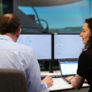 Two people seated at a desk viewing multiple computer screens, working on digital systems in a modern office or control room environment.