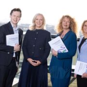 Four business leaders stand side by side on a rooftop terrace overlooking a city, each holding a copy of the Open Doors Initiative report on disability employment.