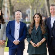 Four people stand outdoors in a park on a sunny day, smiling at the camera. From left to right: a woman in a light grey blazer and white trousers, a man in a blue suit, a woman in a dark green suit, and a man in a dark navy suit. Trees and a pathway are visible in the background.