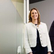 A professional stands in a modern office environment, wearing a light-coloured structured jacket over dark clothing. The setting includes glass walls and contemporary interior design elements, with soft lighting and abstract office furnishings visible in the background.