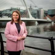 Woman in a pink blazer standing by a riverside railing in Dublin, with a modern pedestrian bridge and city buildings in the background.