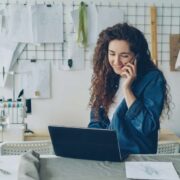 Small business owner working at a desk with a laptop, taking a phone call in a design studio with sketches, fabric, and clothing samples in the background.
