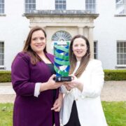 Two people standing outdoors in front of a Georgian university building, holding a glass and metal NovaUCD award trophy together, with a lawn and building fa&ccedil;ade visible in the background.