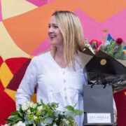 Person wearing a white buttoned top stands holding two bouquets of flowers and a black gift bag labeled “LULABELLE FLOWERS,” positioned in front of a brightly coloured abstract mural wall.