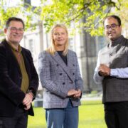 Three people standing outdoors on a college campus lawn with historic stone buildings behind them, dressed in business attire, facing the camera in natural daylight.