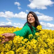 Raquel Noboa, founder of Fifty Shades Greener, smiling as she reaches out across a field of bright yellow flowers under a blue, cloud-filled sky.