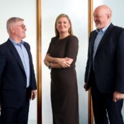 Three business professionals standing indoors against a light backdrop, wearing formal business attire, positioned side by side and facing slightly toward one another.