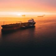 Oil tanker at sea during sunset with several cargo ships on the horizon, glowing orange light reflecting on calm water amid global energy supply concerns.