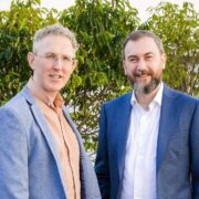 Two business executives standing outdoors in front of greenery, wearing smart jackets and shirts, posed side by side for a professional photograph.