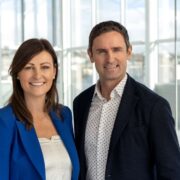 Two people standing side by side in a bright modern office space with large glass windows, wearing business attire and facing the camera.