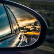 Car side mirror reflecting a winding road and dramatic sunset sky, suggesting travel disruption and uncertainty on the journey ahead.